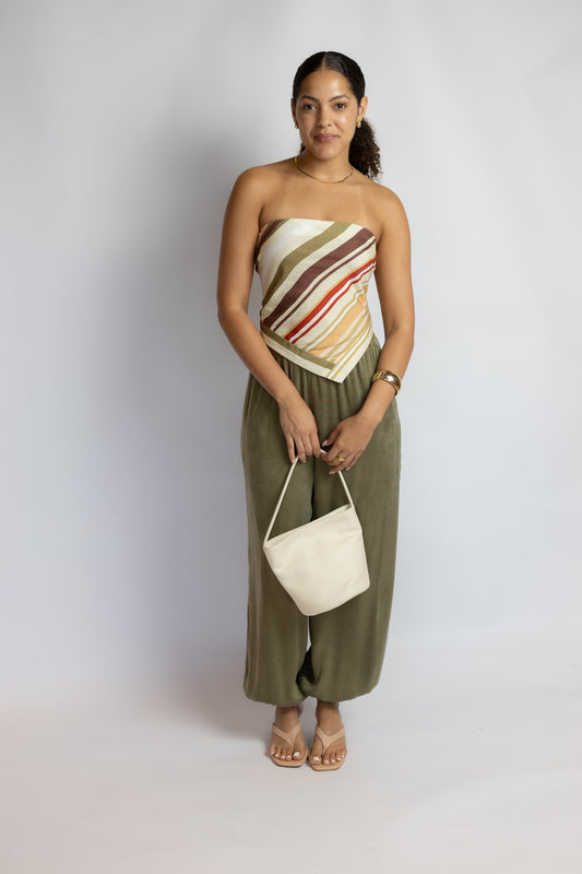 A woman stands against a light gray studio background wearing a strapless two-piece outfit with a diagonal striped top in beige, brown, and red tones, olive green wide-leg pants, and holding a small off-white shoulder bag.