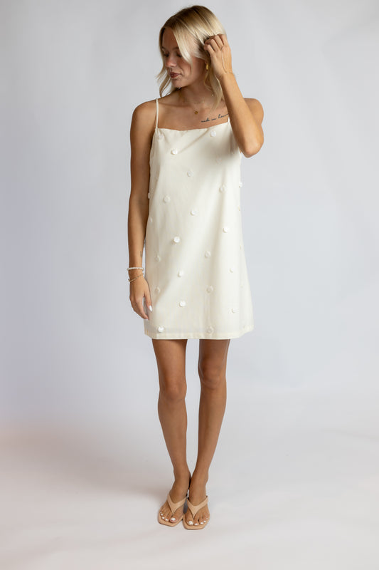 A woman modeling a short cream-colored spaghetti-strap dress with 3D pom-pom details, paired with beige flat sandals, against a light gray studio background.