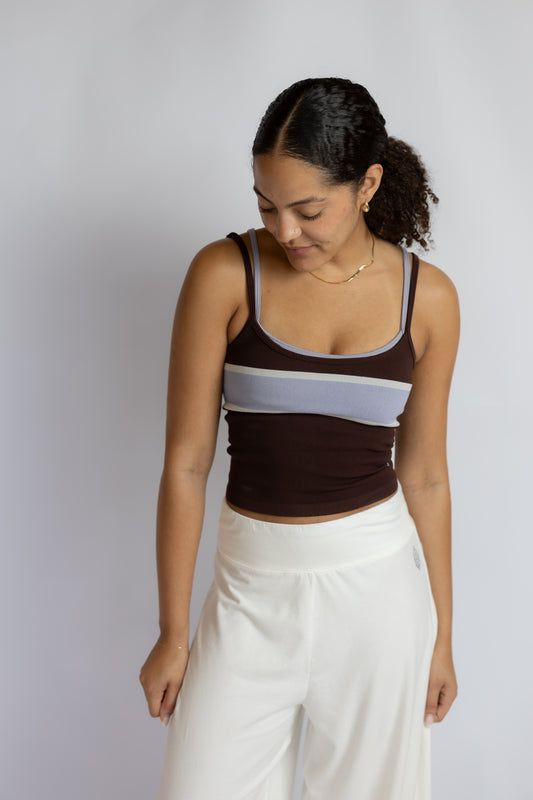 A woman wearing a dark brown ribbed tank top with a light gray horizontal stripe across the chest and white pants, standing against a light gray studio background.