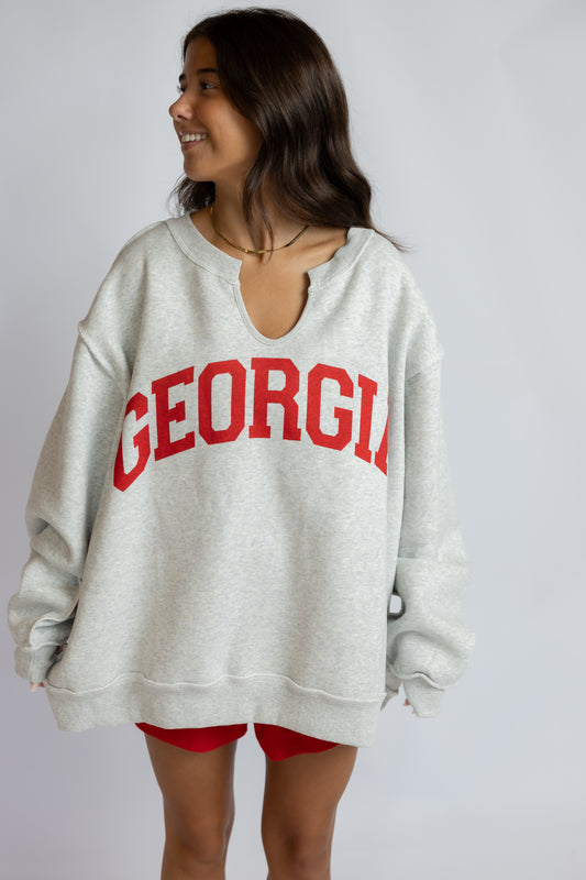 A woman wearing an oversized light gray heather sweatshirt with 'GEORGIA' printed in red arched letters across the chest, paired with red shorts, against a plain light gray background.