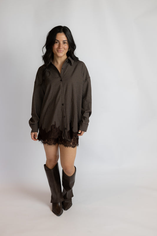 A woman modeling a dark brown and black checkered button-down shirt with a lace hem, paired with dark brown knee-high boots, against a plain light gray studio background.