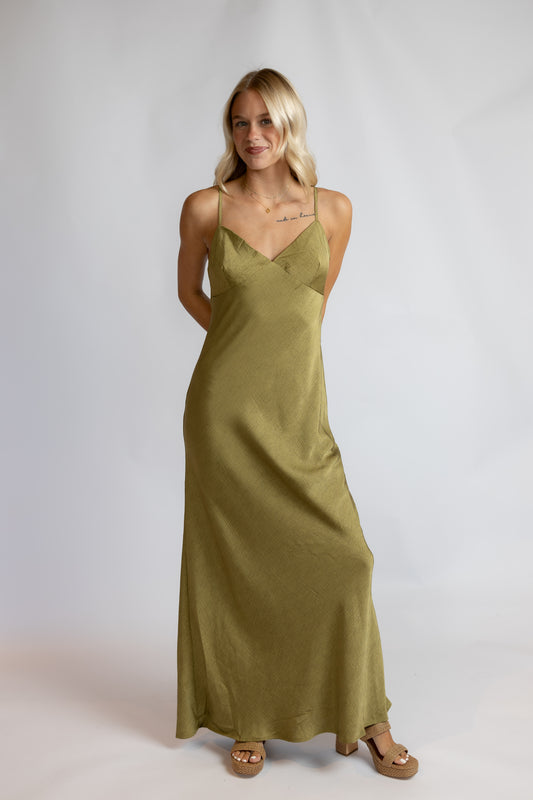A woman modeling a long olive green satin slip dress with spaghetti straps and a V-neckline, paired with brown sandals, against a light gray studio background.