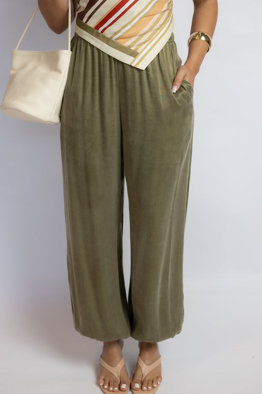 A person wearing olive green wide-leg cropped pants with a striped top and beige sandals, holding a cream-colored shoulder bag against a light gray studio background.