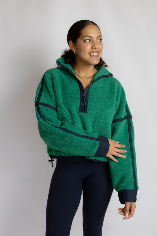 A woman modeling a green fleece half-zip pullover with dark navy contrast panels and trim, paired with dark leggings, against a plain light gray studio background.