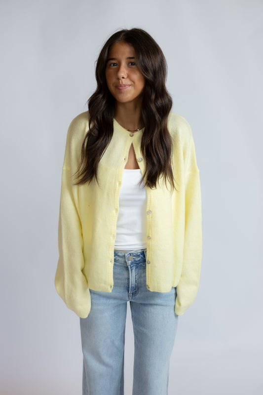 A woman modeling a pale yellow knit cardigan over a white tank top and light blue jeans, standing against a plain light gray studio background.