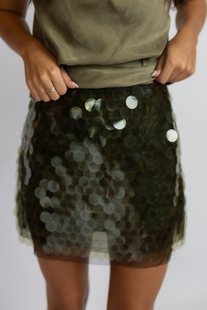 A close-up of a person wearing a short, sheer tulle mini skirt covered in dark olive green sequins with scattered iridescent white and silver discs, paired with an olive green top against a light gray background.