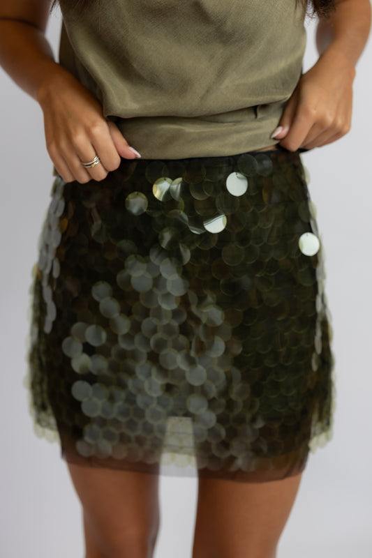 A close-up of a person wearing a short, sheer tulle mini skirt covered in dark olive green sequins with scattered iridescent white and silver discs, paired with an olive green top against a light gray background.