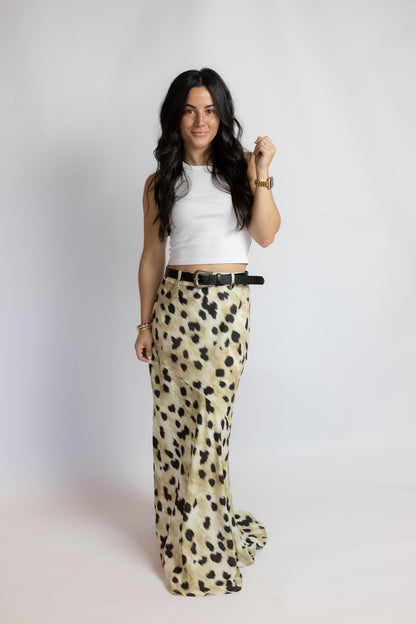 A woman modeling a pair of wide-leg trousers in a beige and black leopard print, paired with a white sleeveless crop top and a black belt, standing against a light gray studio background.