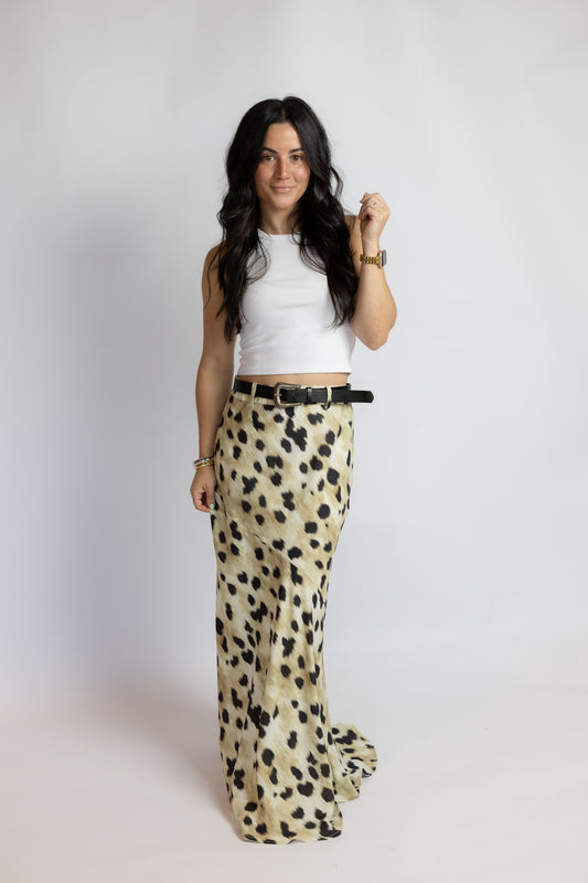 A woman modeling a pair of wide-leg trousers in a beige and black leopard print, paired with a white sleeveless crop top and a black belt, standing against a light gray studio background.
