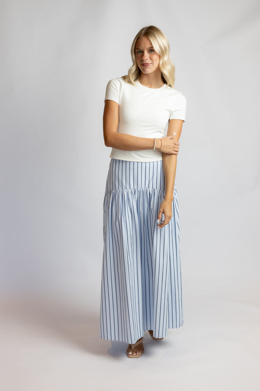 A woman modeling a white short-sleeve top and a high-waisted, wide-leg skirt with vertical light blue and white stripes, standing against a light gray studio backdrop.