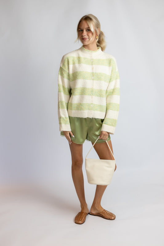 A woman stands against a light gray studio background wearing a fuzzy off-white and pale green striped mock-neck sweater, olive green satin shorts, tan woven mule sandals, and holding a cream canvas bucket bag.