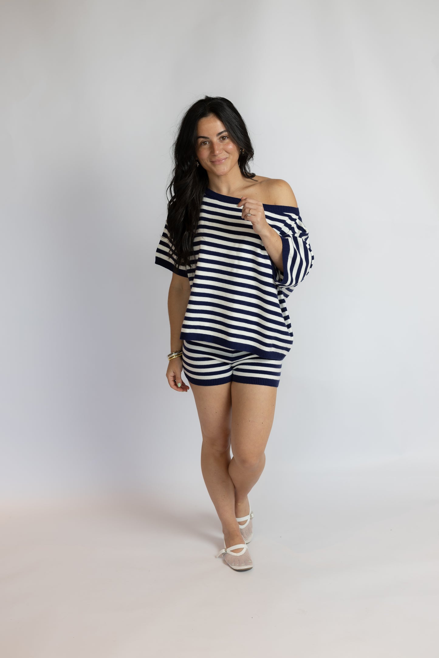 A woman modeling a matching navy and white striped off-the-shoulder t-shirt and shorts set, standing against a light gray studio backdrop.