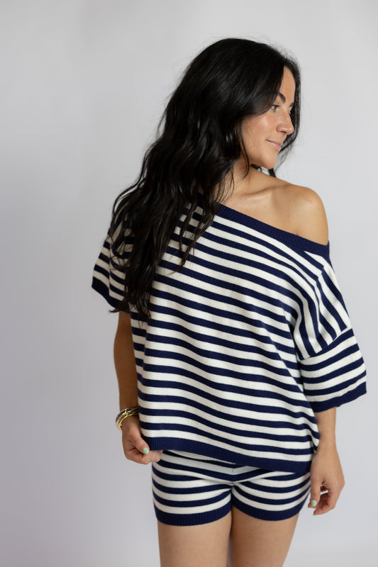 A woman wearing a navy blue and white horizontally striped off-the-shoulder top and matching striped shorts, photographed against a plain light gray studio background.