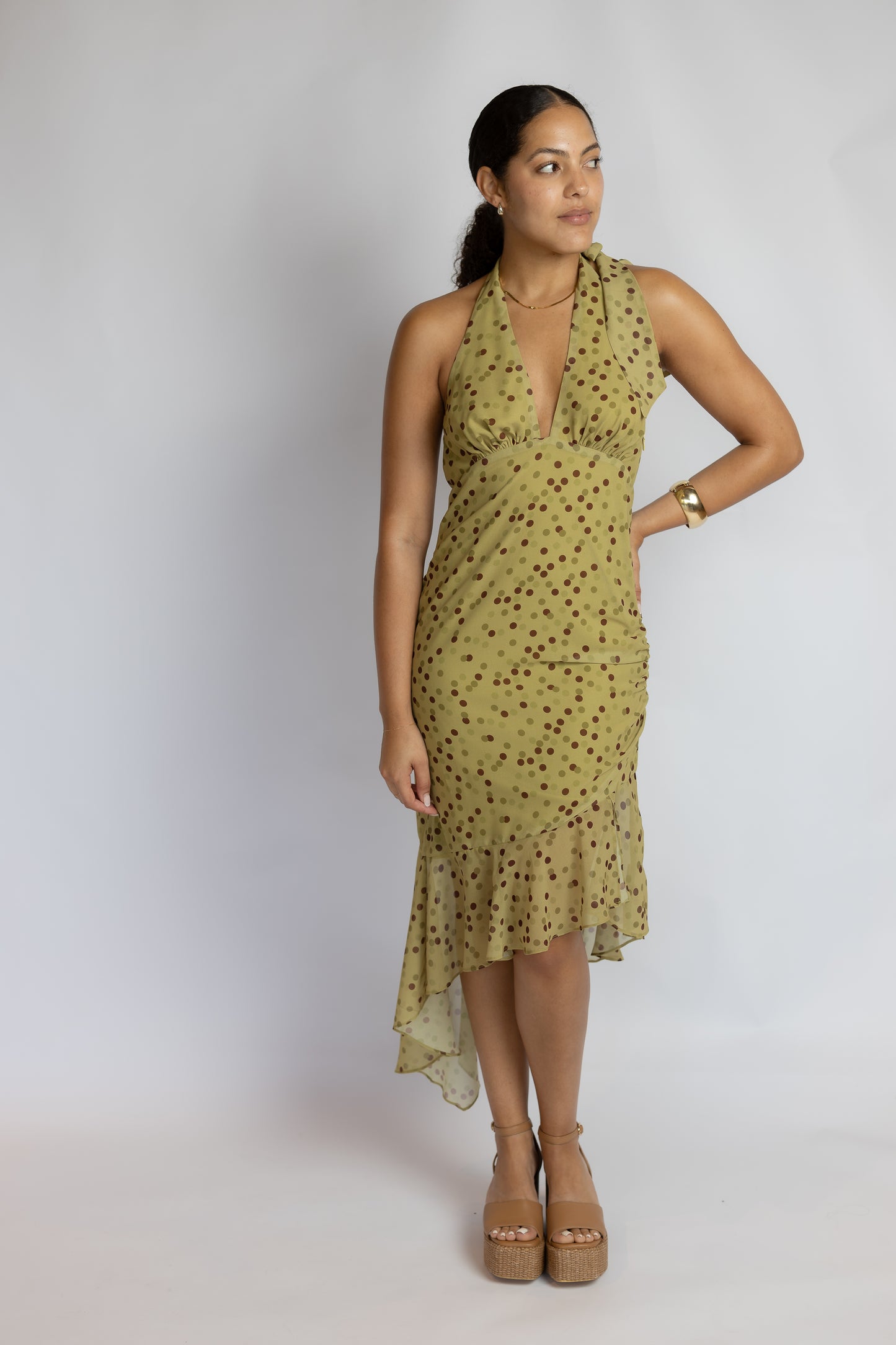 A woman modeling a sleeveless olive green halter-neck dress with a dark brown polka dot pattern, standing against a light gray studio background.