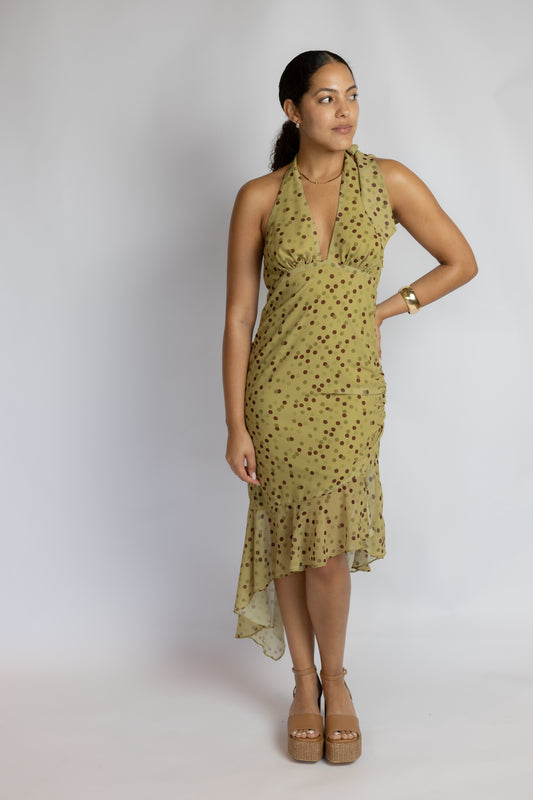 A woman modeling a sleeveless olive green halter-neck dress with a dark brown polka dot pattern, standing against a light gray studio background.