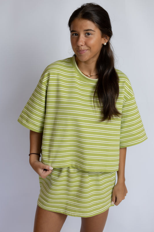 A young girl with long dark hair wearing a lime green and white striped lounge set, consisting of a short-sleeve t-shirt and a matching mini skirt, against a plain light gray studio background.