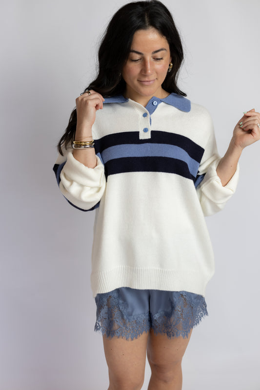 A woman modeling a knit polo-style sweater with a cream base and horizontal navy and dusty blue stripes, paired with blue lace shorts, against a light gray studio background.
