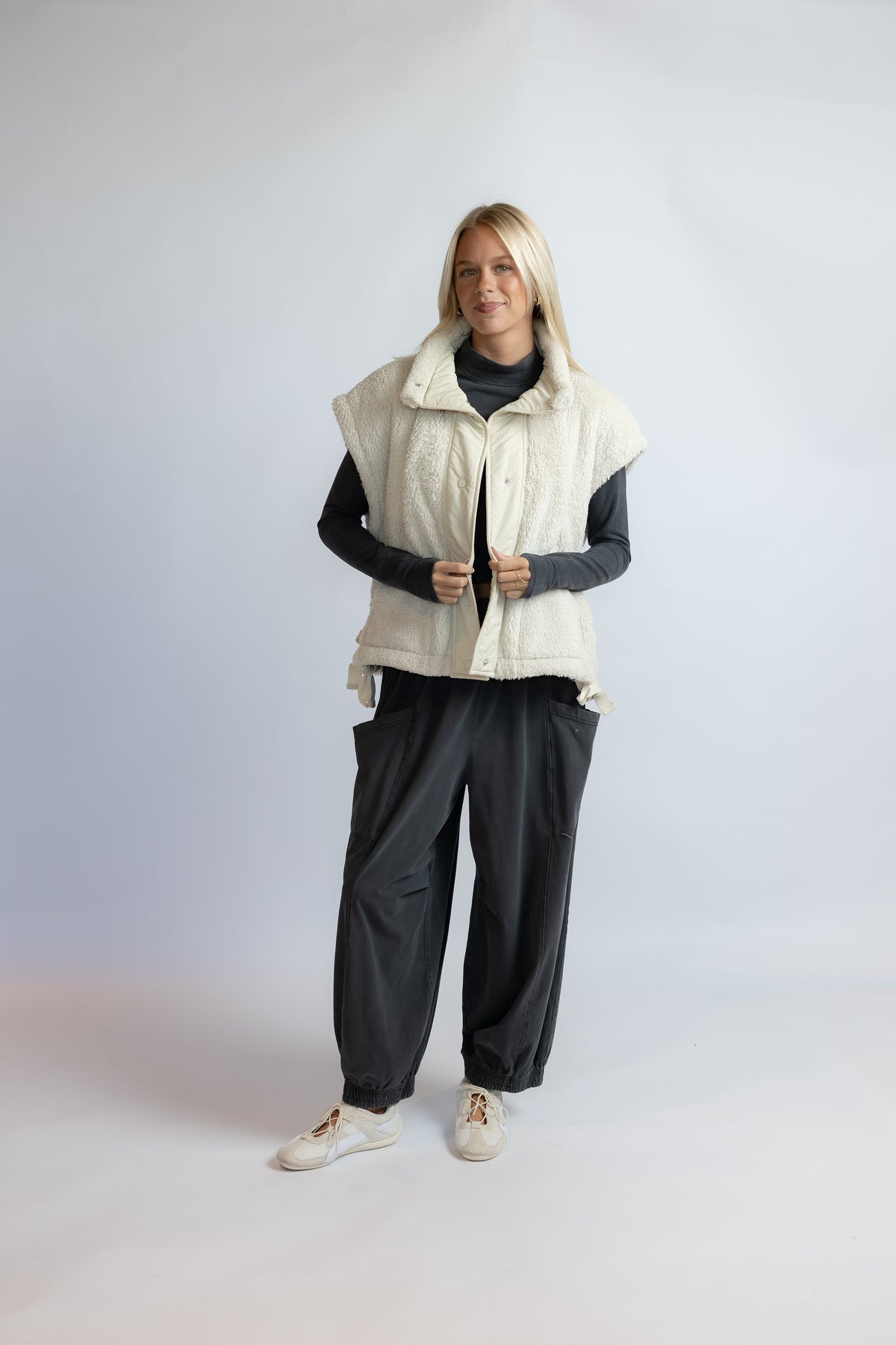 A woman modeling a cream-colored sleeveless quilted vest over a black long-sleeve top, black wide-leg pants, and white sneakers, standing against a light gray studio background.