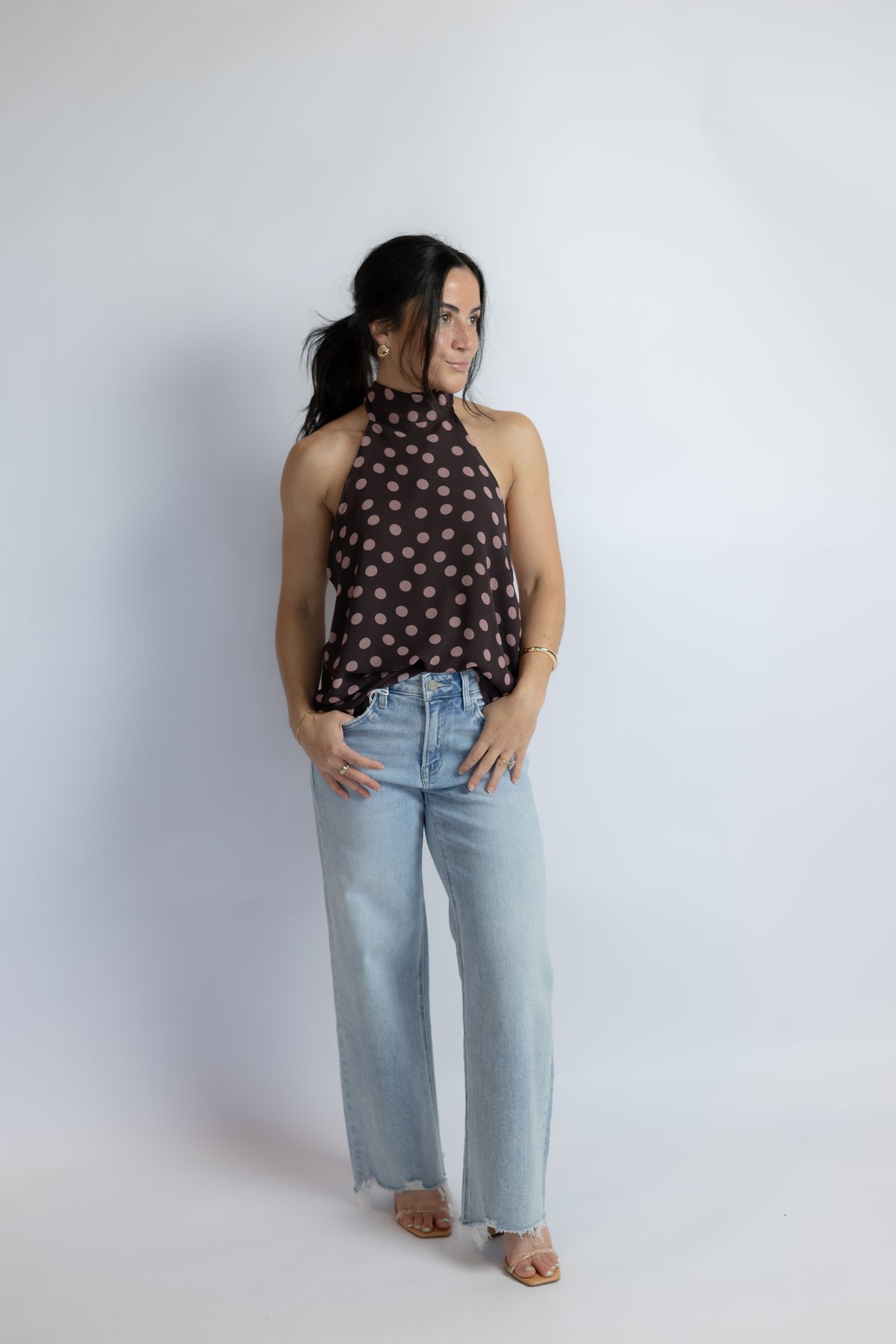 A woman modeling a dark brown halter top with a beige polka dot pattern, paired with light-wash wide-leg jeans, standing against a light gray studio background.