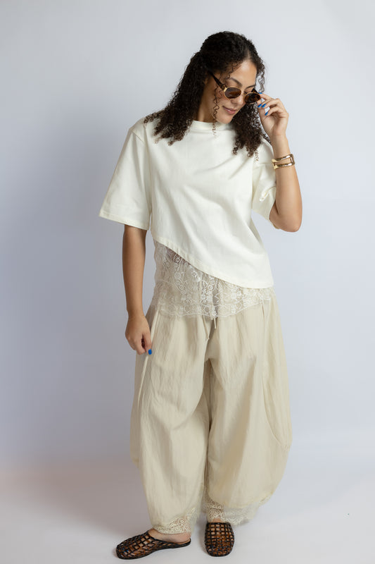 A woman modeling a cream-colored outfit consisting of a short-sleeve asymmetrical top, a lace-trimmed beige skirt or drape, wide-leg beige pants, and brown woven sandals, set against a plain light gray studio background.