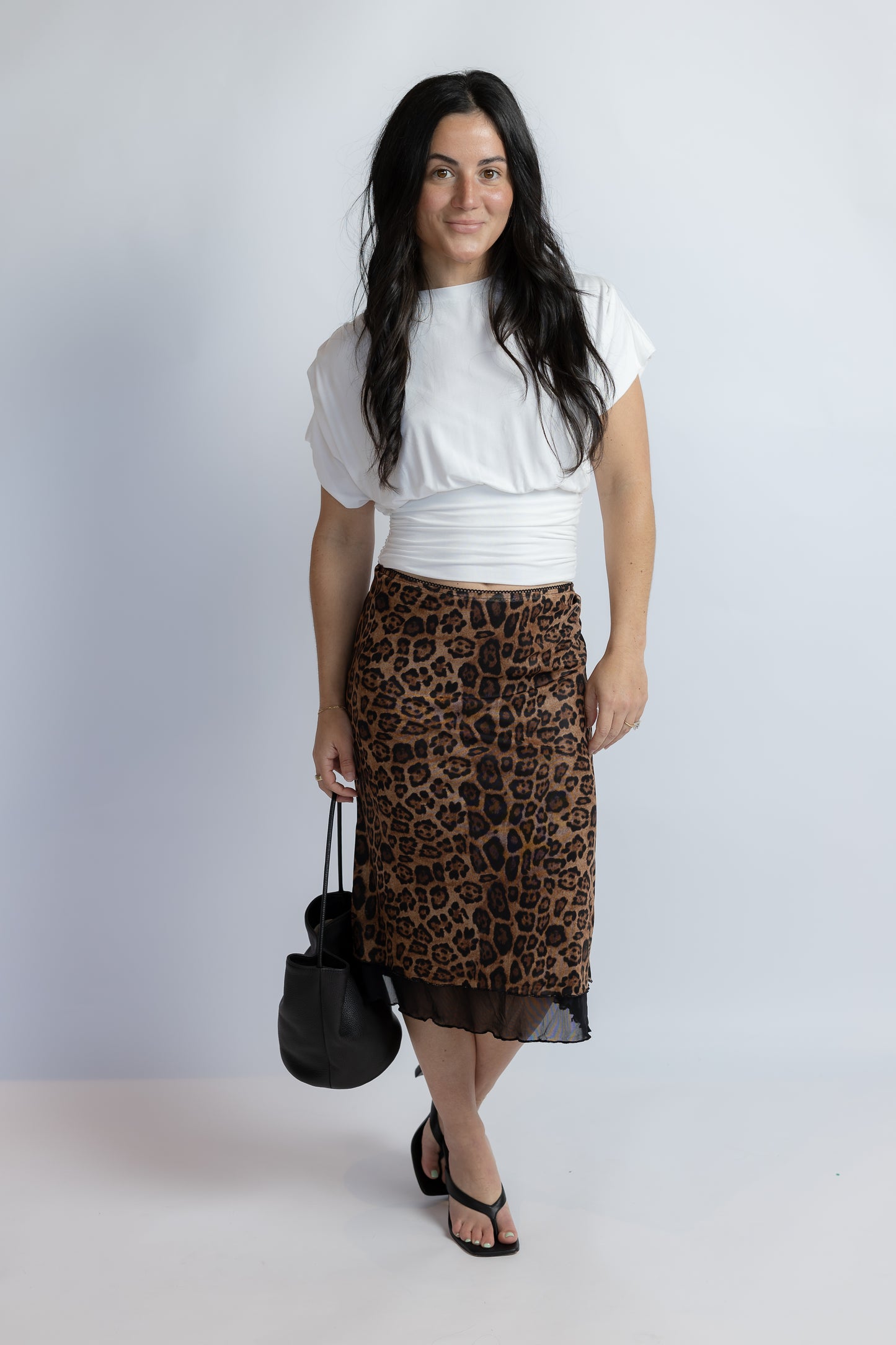 A woman modeling a brown and black leopard print midi skirt with a sheer black hem, styled with a white cropped top, black sandals, and a black handbag against a light gray studio background.