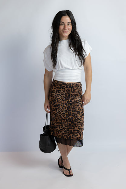 A woman modeling a brown and black leopard print midi skirt with a sheer black hem, styled with a white cropped top, black sandals, and a black handbag against a light gray studio background.
