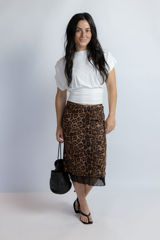 A woman modeling a brown and black leopard print midi skirt with a sheer black hem, styled with a white cropped top, black sandals, and a black handbag against a light gray studio background.
