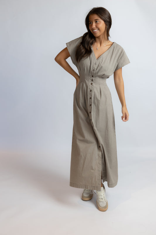 A woman modeling a taupe linen midi dress with short sleeves, a V-neckline, and a button-down front, paired with white sneakers, against a light gray studio background.