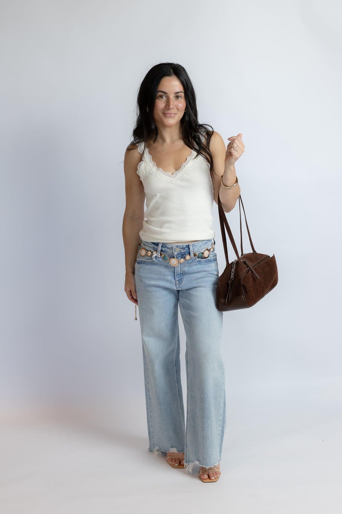 A woman modeling a white lace-trimmed camisole top, light-wash wide-leg jeans with a decorative gold chain belt, and carrying a brown leather shoulder bag against a light gray studio background.