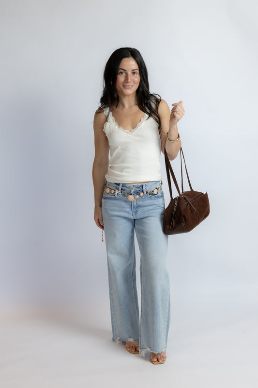 A woman modeling a white lace-trimmed camisole top, light-wash wide-leg jeans with a decorative gold chain belt, and carrying a brown leather shoulder bag against a light gray studio background.
