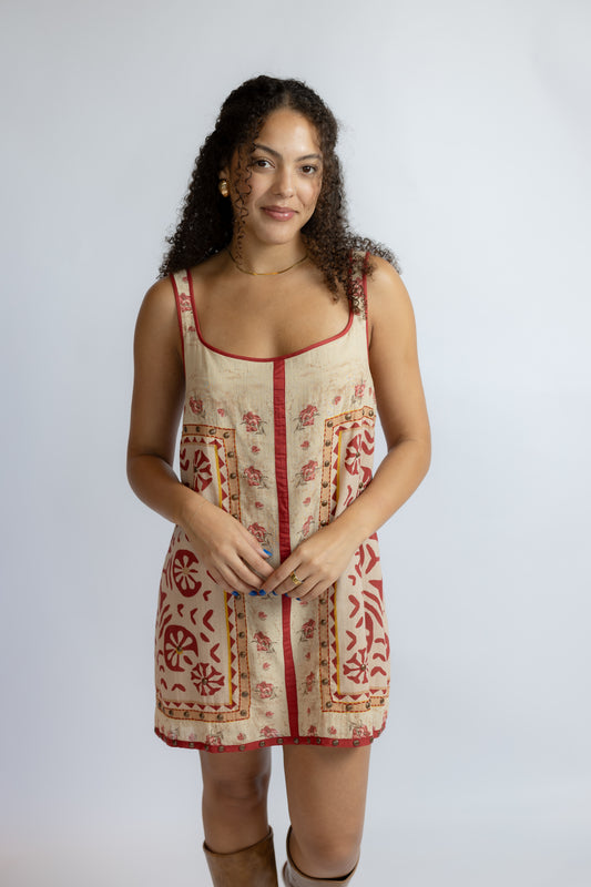 A woman modeling a sleeveless beige linen dress with red and brown bohemian-style patterns and a red center placket, photographed against a light gray studio background.