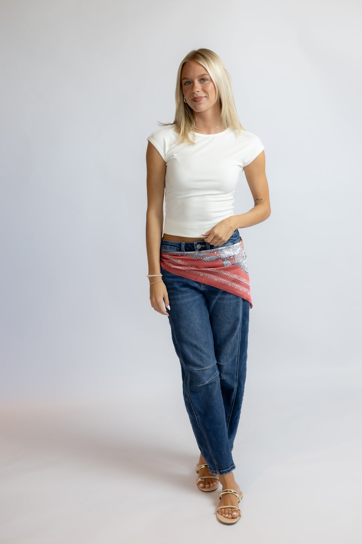 A woman modeling a white short-sleeve crop top, blue jeans, and a red patterned sarong tied around her waist, standing against a light gray studio background.