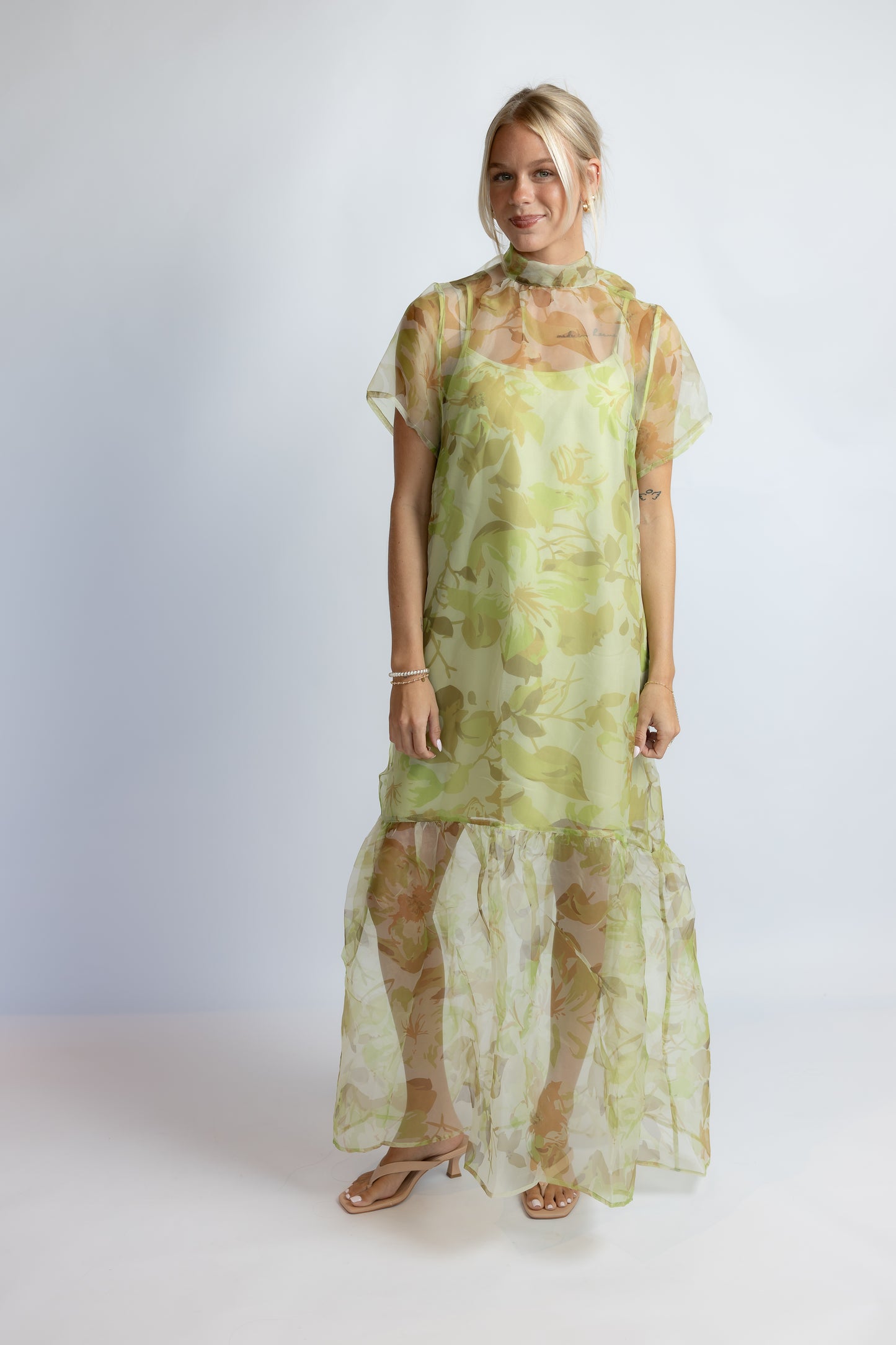 A woman modeling a sheer, light green and yellow floral print maxi dress with short flutter sleeves and a high neck, paired with tan flat sandals, against a light gray studio background.