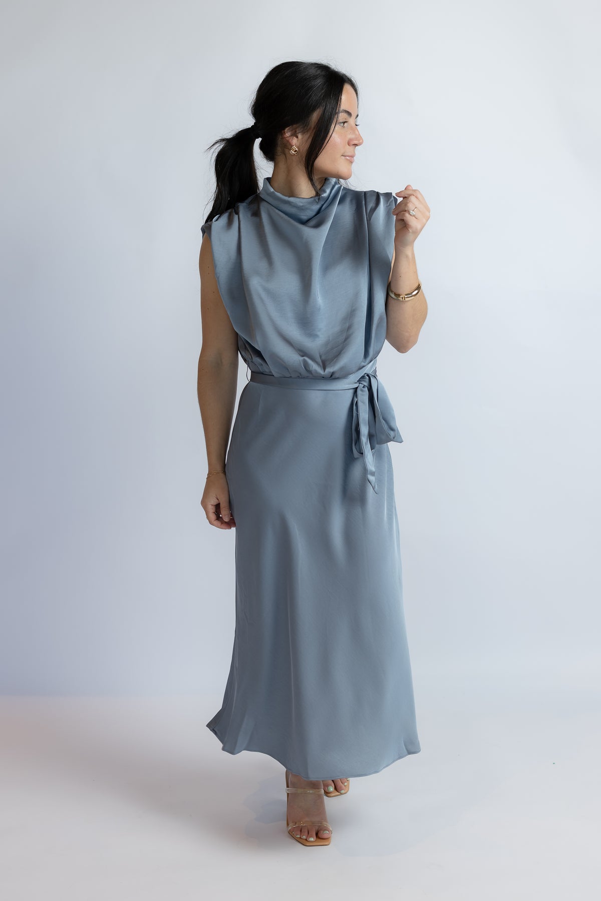 A woman modeling a sleeveless, high-neck, cowl-neck maxi dress in a dusty slate blue color, featuring a tie-waist belt and flowing skirt, set against a plain light gray studio background.