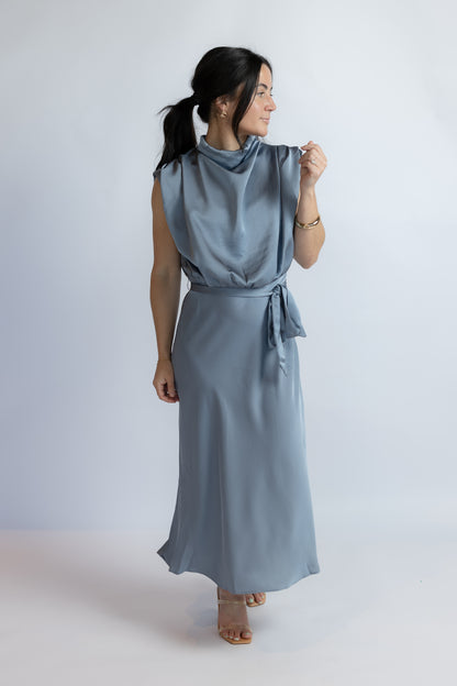 A woman modeling a sleeveless, high-neck, cowl-neck maxi dress in a dusty slate blue color, featuring a tie-waist belt and flowing skirt, set against a plain light gray studio background.
