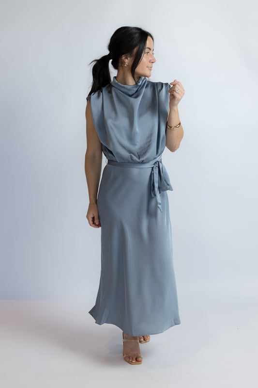 A woman modeling a sleeveless, high-neck, cowl-neck maxi dress in a dusty slate blue color, featuring a tie-waist belt and flowing skirt, set against a plain light gray studio background.