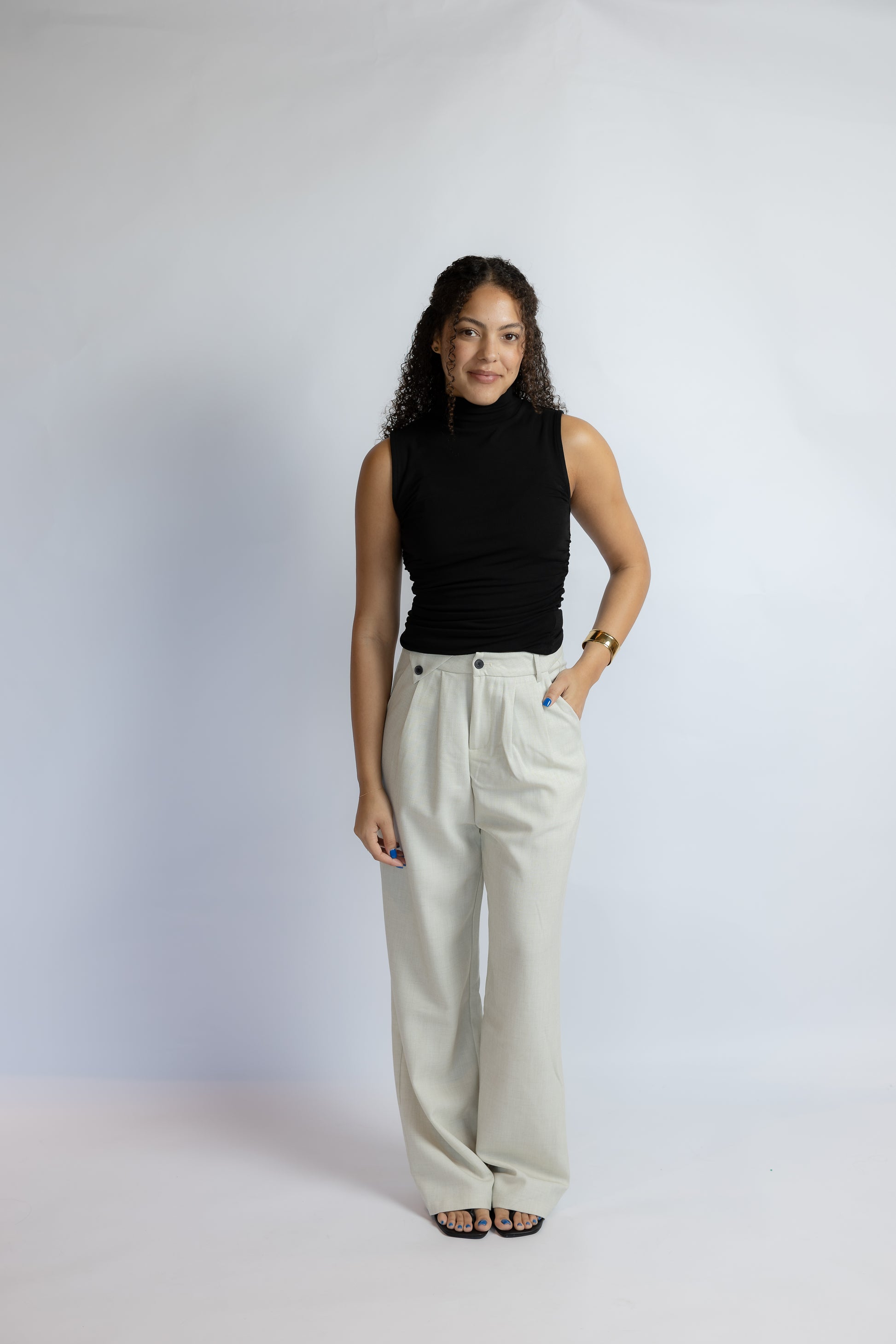A woman modeling a black sleeveless mock neck top and high-waisted, wide-leg light beige trousers against a light gray studio backdrop.