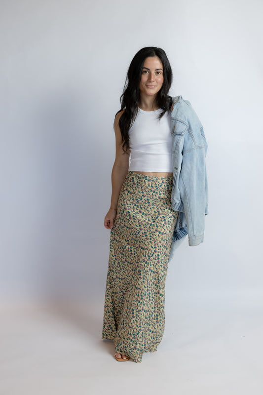 A woman modeling a long, wide-leg maxi skirt with a green and blue floral print on a beige background, styled with a white crop top and a light blue denim jacket draped over her shoulder.
