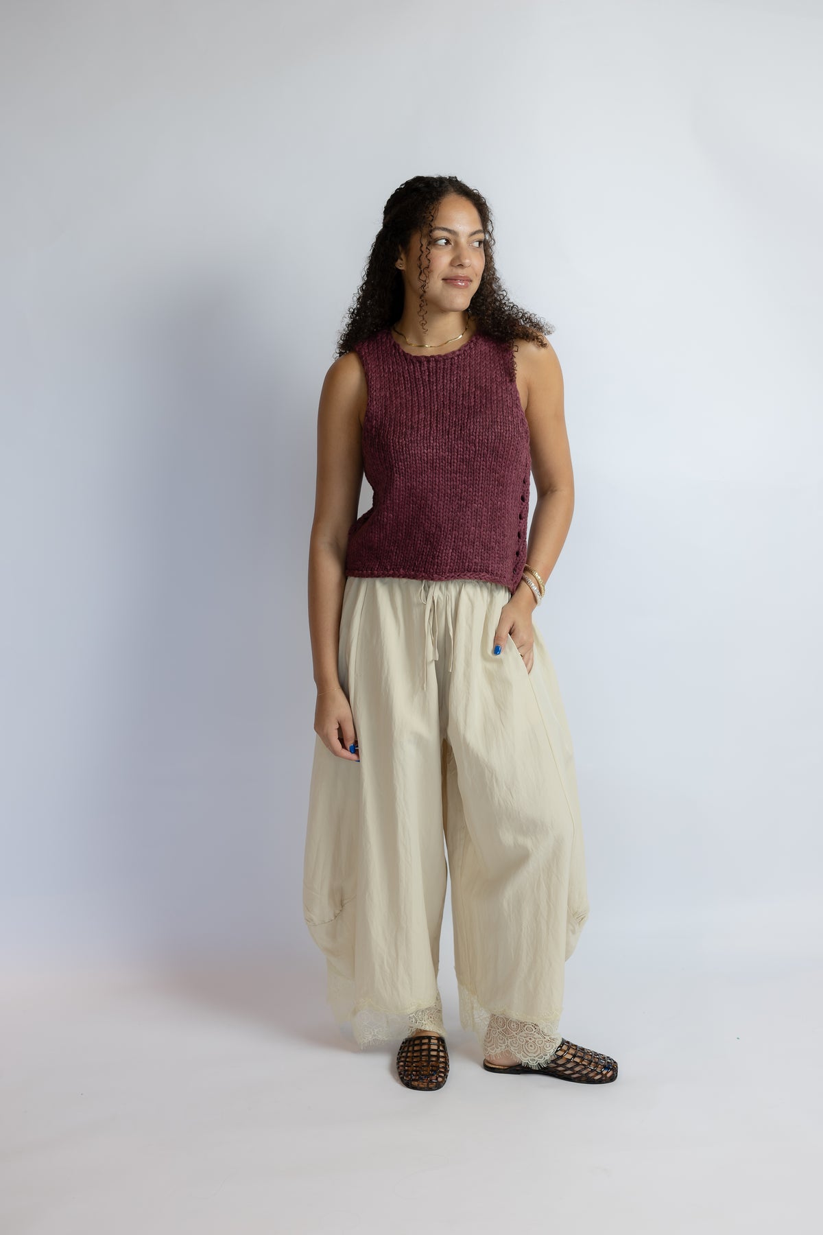 A woman modeling a sleeveless maroon knit top and cream wide-leg pants against a light gray studio background.