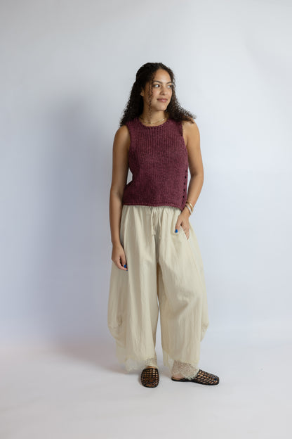 A woman modeling a sleeveless maroon knit top and cream wide-leg pants against a light gray studio background.