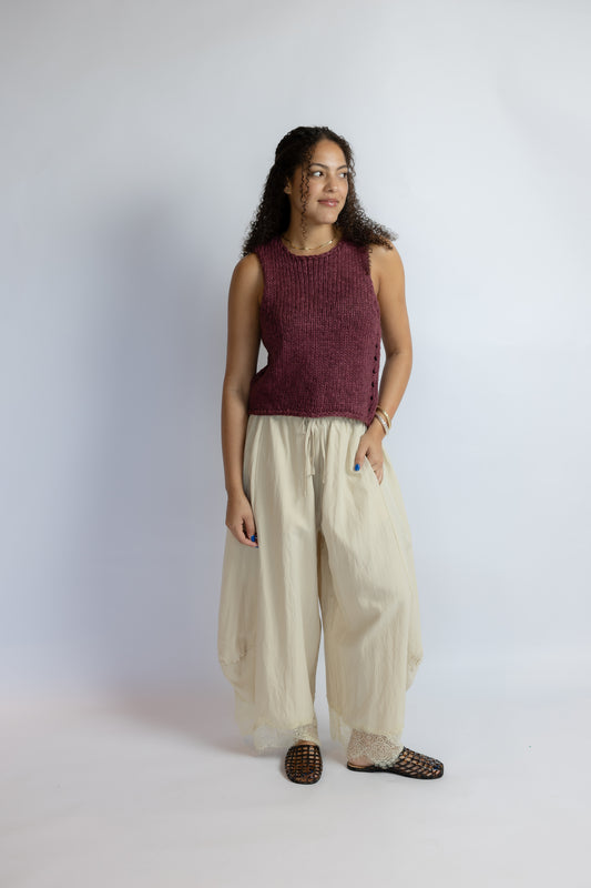 A woman modeling a sleeveless maroon knit top and cream wide-leg pants against a light gray studio background.