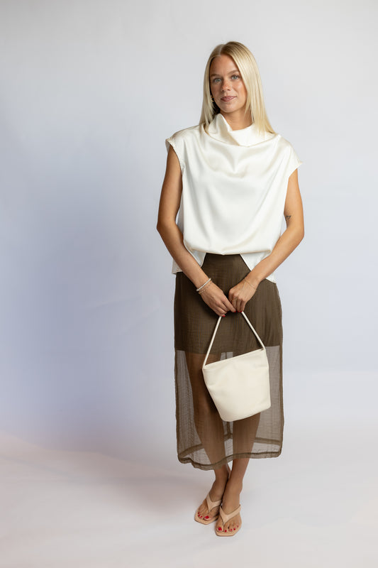 A blonde woman stands against a light gray studio background wearing a white sleeveless cowl-neck top, a brown midi skirt with a sheer overlay, and holding a white bucket bag.