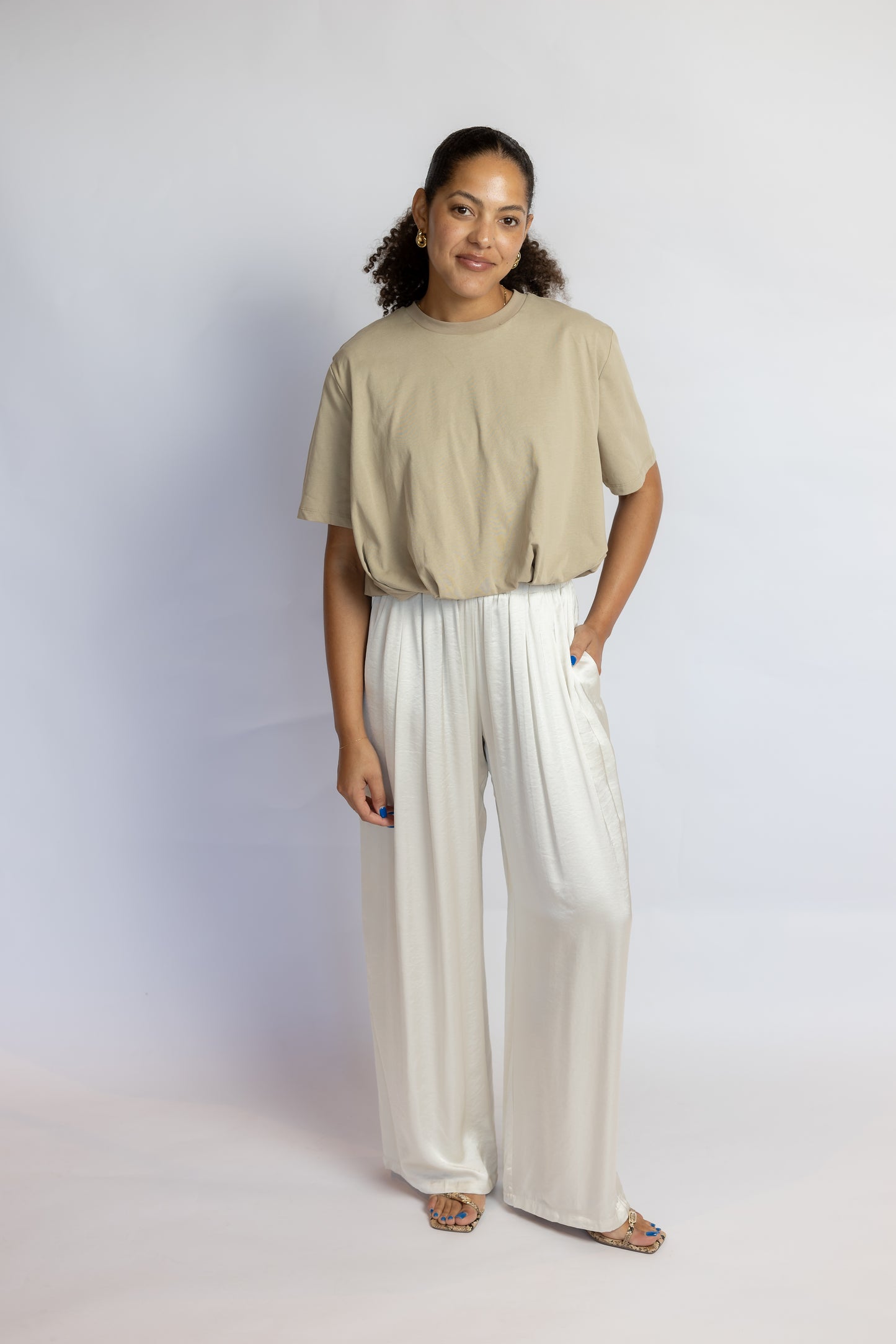 A woman modeling a beige short-sleeve crop top and white wide-leg pants against a light gray studio background.