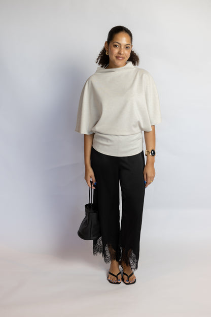 A woman modeling a cream-colored short-sleeve knit top and black high-low pants with lace trim, holding a black handbag against a light gray studio background.