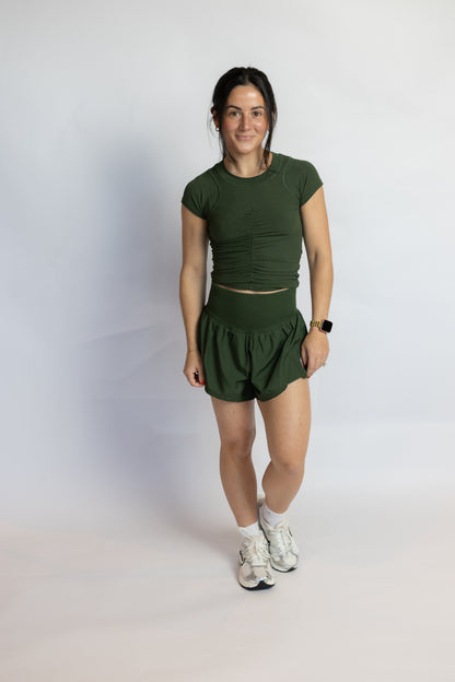 A woman modeling an olive green athletic set consisting of a short-sleeve crop top and matching high-waisted shorts, paired with white socks and running shoes, against a plain light gray studio background.