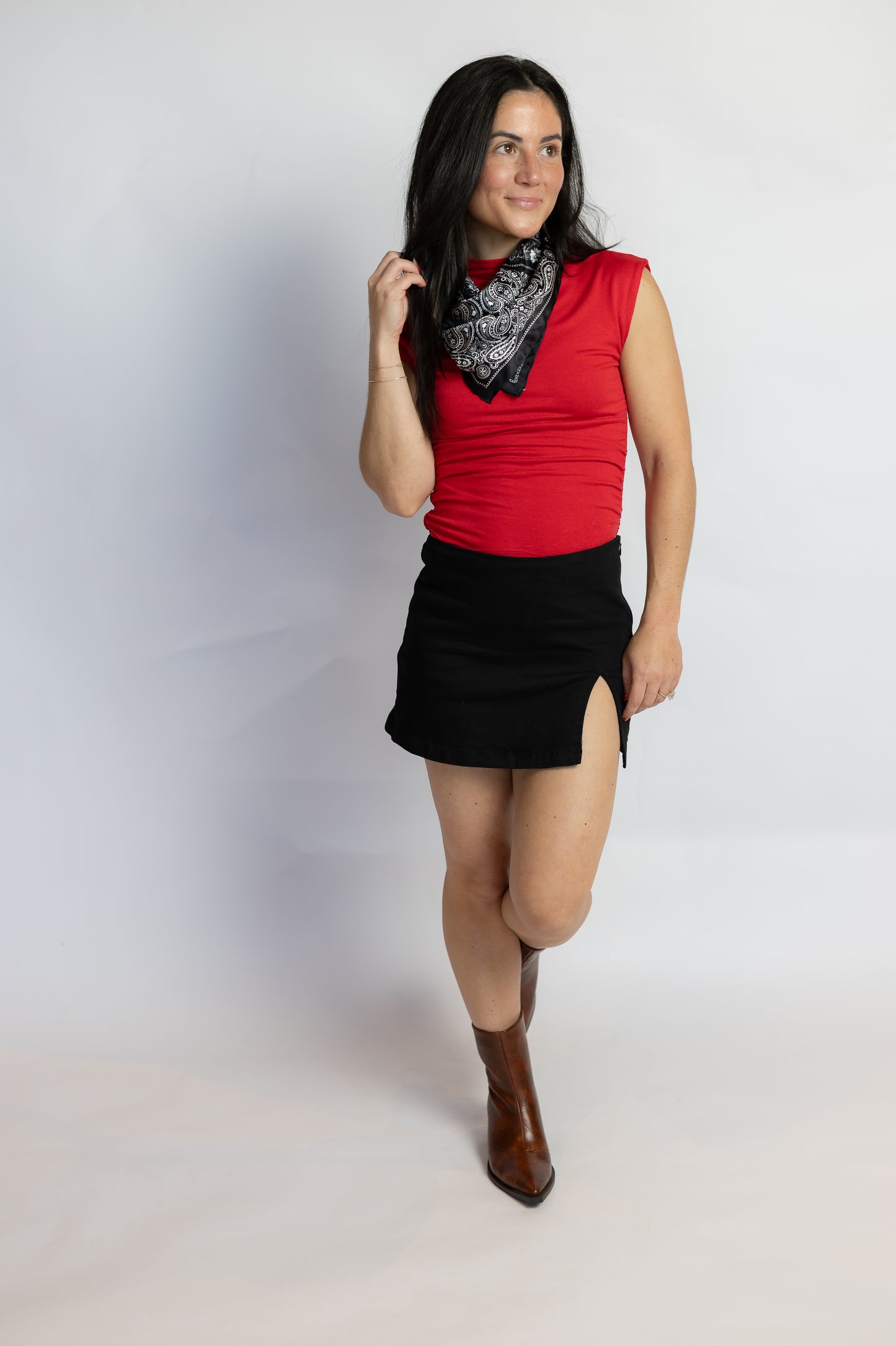 A woman modeling a sleeveless red top, black mini skirt with side slits, a black and white patterned bandana, and brown ankle boots, standing against a light gray studio background.