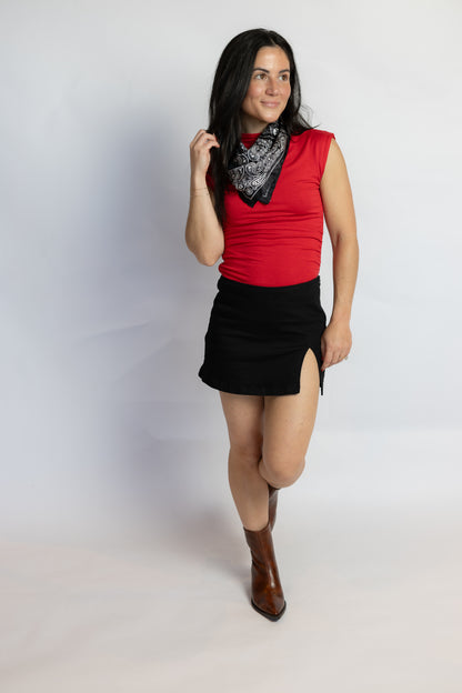A woman modeling a sleeveless red top, black mini skirt with side slits, a black and white patterned bandana, and brown ankle boots, standing against a light gray studio background.