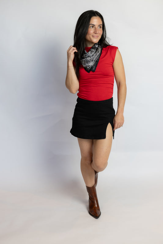 A woman modeling a sleeveless red top, black mini skirt with side slits, a black and white patterned bandana, and brown ankle boots, standing against a light gray studio background.