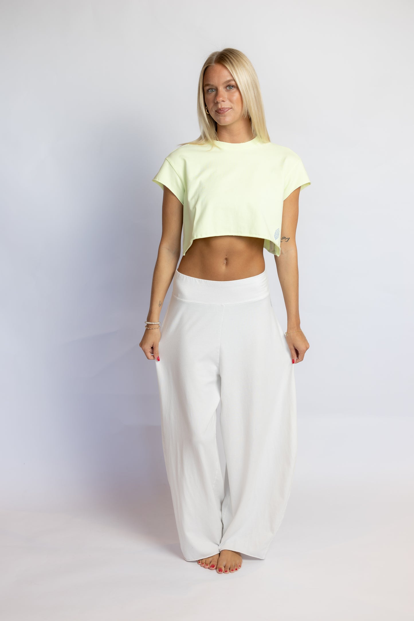 A blonde woman stands barefoot against a light gray studio background, wearing a pale yellow-green short-sleeve cropped t-shirt and loose white wide-leg pants.
