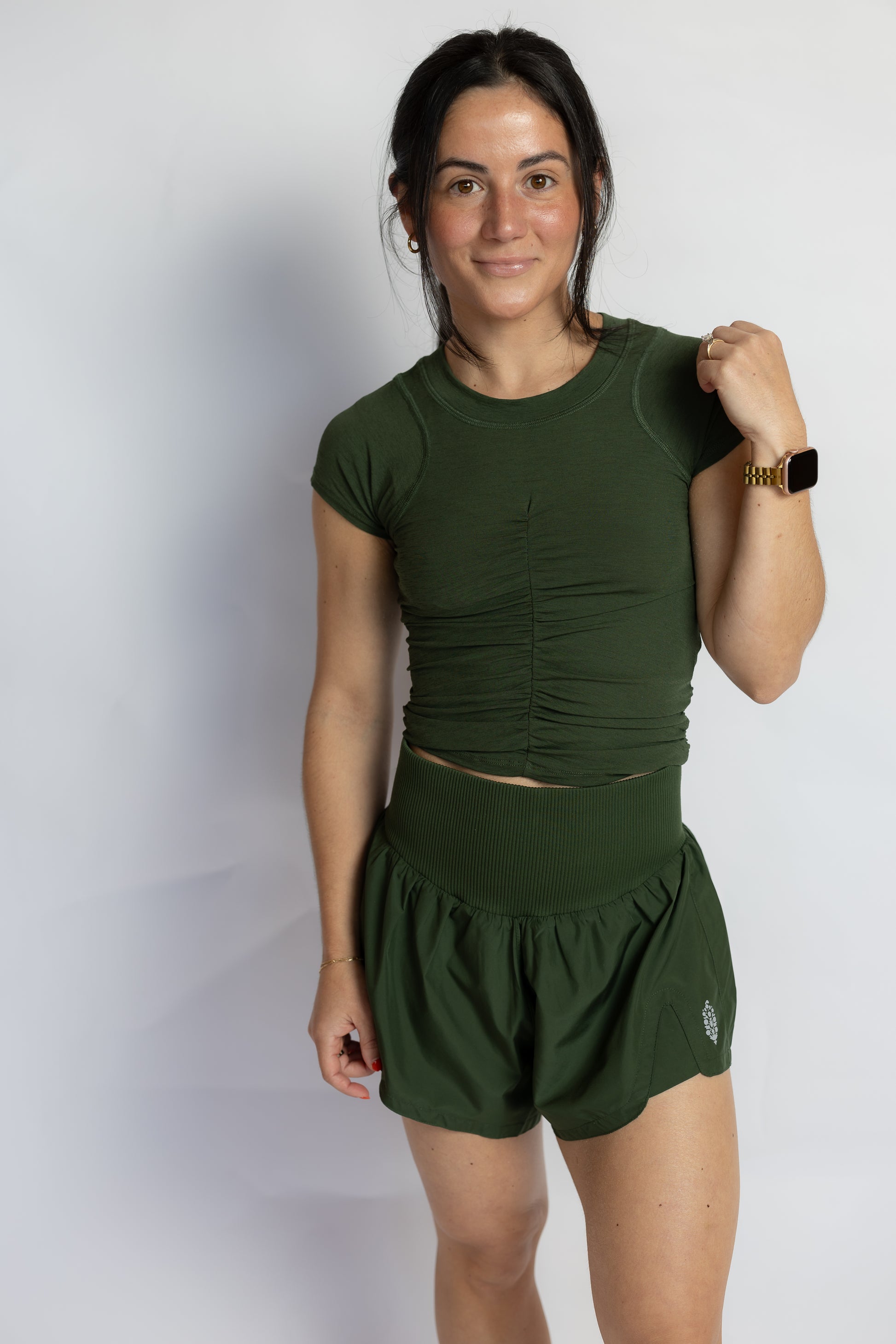 A woman modeling a matching dark green athletic set consisting of a short-sleeve ruched crop top and high-waisted shorts, standing against a plain light gray background.